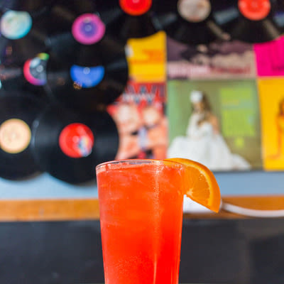 a bright orange-red cocktail in a pint glass with an orange garnish with records in the background