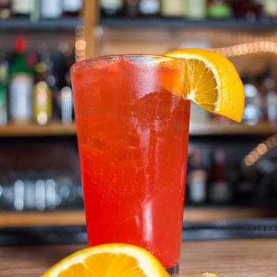 a bright orange-red cocktail in a pint glass with an orange garnish