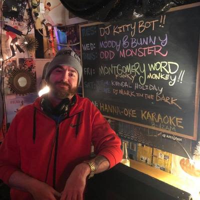 a male DJ posed in the DJ booth, known as the Cat Box, next to the chalkboard DJ schedule