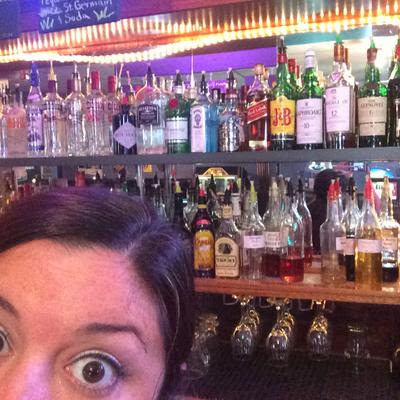 A Mad Hanna bartender taking a selfie of the top half of her face with call and premium bottles of alcohol in the background