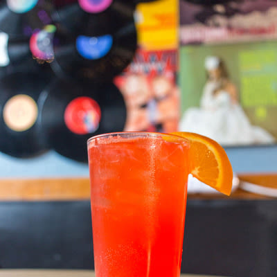 a bright orange-red cocktail in a pint glass with an orange garnish with records in the background